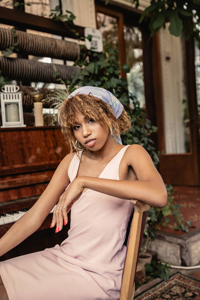 Retrato de jovem e elegante mulher afro-americana em vestido olhando para a câmera enquanto sentado perto de piano e plantas em jardim interior borrado, mulher em roupa de verão confortável e na moda - Foto, Imagem