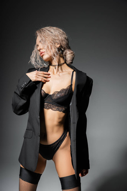 fashion-forward and sexy woman in black blazer, panties and lace bra posing on grey background, dyed and wavy ash blonde hair, representation of stylish lingerie on slender body - Photo, Image