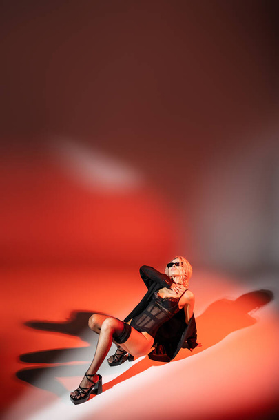 сексуальність і мода, повна довжина провокаційної жінки в темних сонцезахисних окулярах, чорний боді-костюм, блейзер, панчохи і каблуки сандалі позують на червоному і сірому тлі з копіювальним простором - Фото, зображення