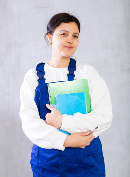 Portrait of positive young dark-haired woman, professional cleaning service worker, wearing blue uniform standing with order books in hands on gray studio background.. - Foto, imagen