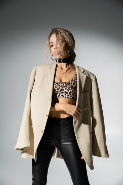 sensuality and fashion, charming woman with dyed ash blonde hair and slender body posing in animal print crop top and black latex pants, with beige blazer on shoulders on grey background - Foto, Bild