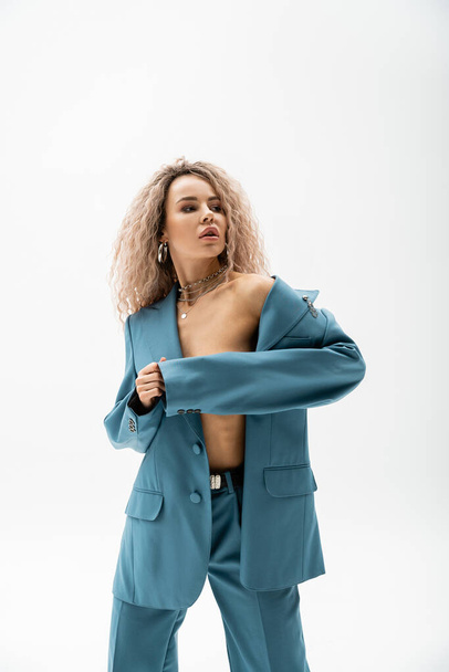 sexuality and fashion, seductive woman with wavy ash blonde hair looking away while wearing blue oversize suit on shirtless body and posing on grey background, expressive individuality - Photo, Image
