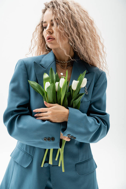 femininity and fashion, appealing and romantic woman with wavy ash blonde hair standing in blue oversize blazer and holding bouquet of white fresh tulips on grey background - Photo, Image