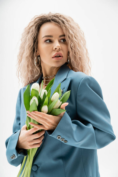 portret modnej kobiety z ekspresyjnym spojrzeniem pozującym z bukietem białych tulipanów i odwracającym wzrok na szarym tle, falujący popiół blond włosy, niebieska marynarka oversize, sesja modowa - Zdjęcie, obraz