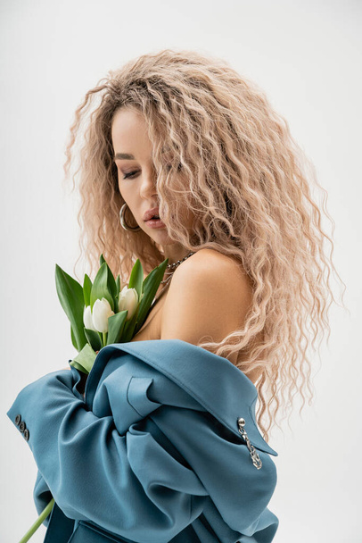 sensual and charming woman with bare shoulders and wavy ash blonde hair holding bouquet of white tulips and posing in blue oversize blazer on grey background, modern fashion and style - Photo, Image