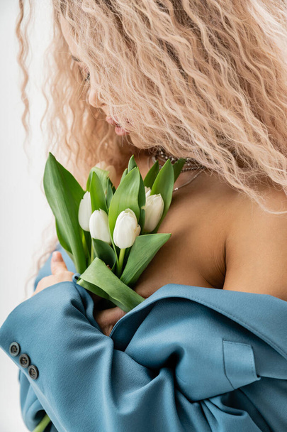 sexuality and fashion, charming and romantic woman with wavy ash blonde hair wearing blue oversize blazer on shirtless body and embracing bouquet of white tulips on grey background - Fotografie, Obrázek