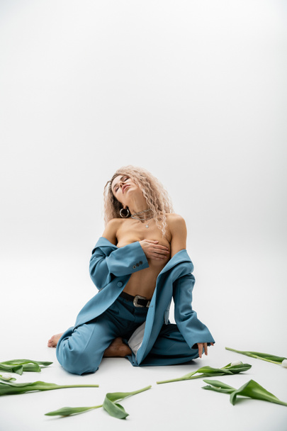 Seksi moda fotoğrafçılığı, boyalı kül sarısı saçlı ve kapalı gözlü tutkulu bir kadın çıplak göğsünü elleriyle örterken gri arka plandaki lalelerin yanında mavi bir takım elbiseyle oturuyor. - Fotoğraf, Görsel