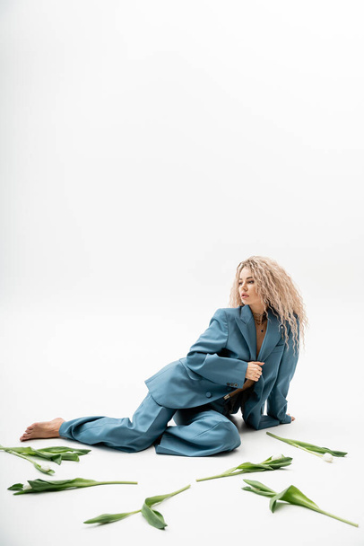 full length of sensual and charming barefoot woman with dyed ash blonde hair wearing blue oversize suit while sitting near tulips on grey background with copy space, modern individuality - Photo, Image