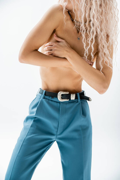 partial view of seductive woman with shirtless body and wavy ash blonde hair obscuring naked breast with hands while posing in silver necklaces and blue pants with leather belt on grey background - Fotografie, Obrázek