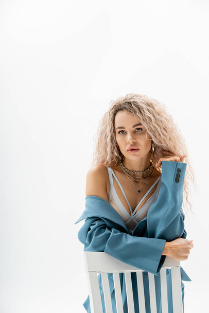 portrait of seductive woman in bra and blue oversize blazer sitting on chair and looking away on grey background, wavy ash blonde hair, expressive gaze, silver accessories, sexy fashion trend - Φωτογραφία, εικόνα