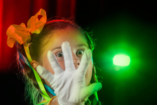 Die kleine Schauspielerin bedeckte ihr Gesicht mit einem weißen Handschuh und blickt durch ihre Finger. Bühnenlicht und Kinderdarsteller. - Foto, Bild