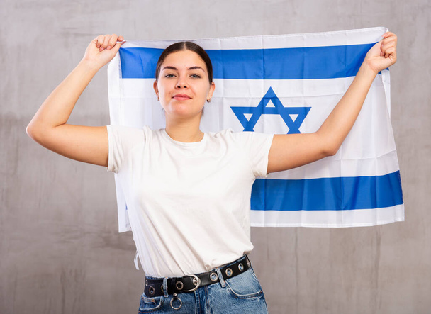 Portrait of serious patriotic young female holding national flag of Israel against gray wall background - Fotografie, Obrázek
