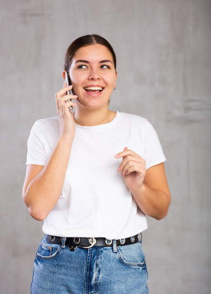 Photo of cheerful young woman speaking on mobile phone with pleasure against gray shadeless background - Foto, Bild