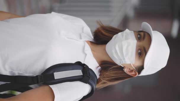 Κάθετη βίντεο. Πορτρέτο της καυκάσιας γυναίκας κούριερ με προστατευτική μάσκα που στέκεται κοντά στο κτίριο. Γρήγορη ασφαλή παράδοση. Ταχυδρομική παράδοση Lady σπίτι. Παράδοση εργαζόμενων γυναικών κατά την πανδημία - Πλάνα, βίντεο