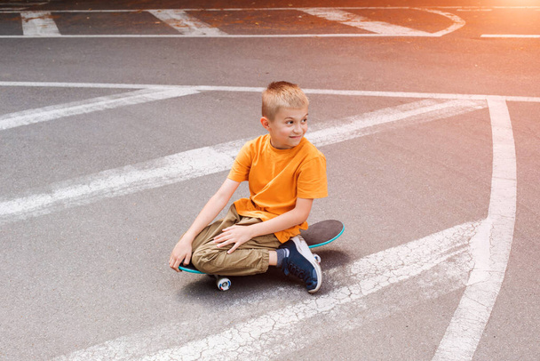 Outdoor-Aktivitäten für Kinder: Ein fröhlicher kleiner Junge in heller Kleidung sitzt auf einem Skateboard. Kaukasischer Schuljunge bei einem Spaziergang. Frontansicht - Foto, Bild