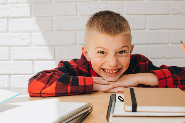 Концепция вернуться в школу. Веселый улыбающийся мальчик в процессе учебы сидит за столом с книгами, блокнотами и карандашами. Вид спереди - Фото, изображение