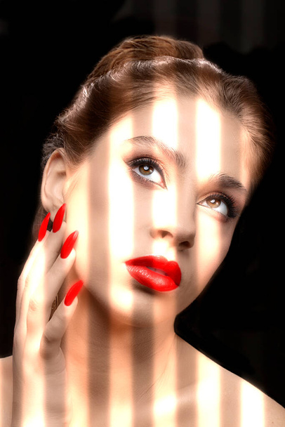 A model with red lipstick and nail polish is illuminated by the bright light of the blinds on a dark background. The face of a beautiful well-groomed girl with clean skin. - Foto, Imagen