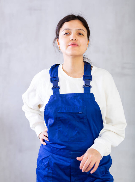 Позитивная молодая женщина, профессиональный строитель, одетая в синий комбинезон позирует уверенно на сером фоне студии, смотрит в камеру с улыбкой - Фото, изображение