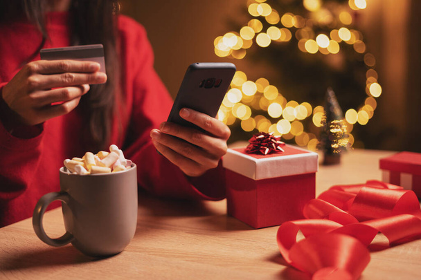 Hispanic woman paying online by credit card for Christmas gifts, close-up hands, using smartphone, sitting at table in the evening, shopping online, enjoying winter holiday. Copy space - Foto, Imagen