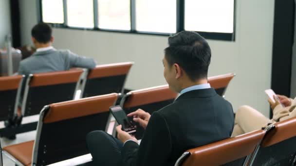 Бизнесмен проверяет информацию о мобильном азиатском бизнесмене, сидящем на стуле в аэропорту, людях, использующих мобильные телефоны для воспроизведения социальных медиа. - Кадры, видео