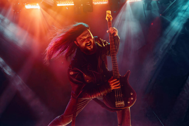 Rock and roll concert. Artistic, expressive man with long hair playing guitar and making performance, emotionally singing. Concept of music, performance, art, talent, nightlife, joy, party, lifestyle - Фото, зображення