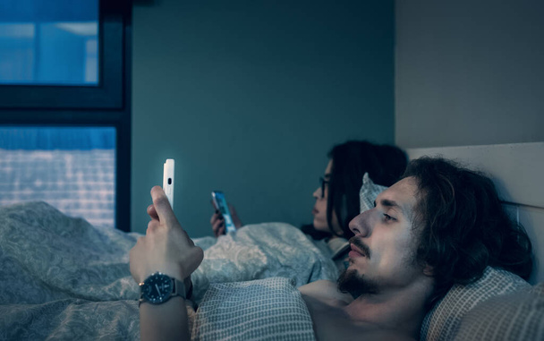 Married couple lying in bed together but each of partners use smartphone. Man and woman looks at mobile phones. Social media addiction, browsing news, problems in relationships. - Фото, изображение