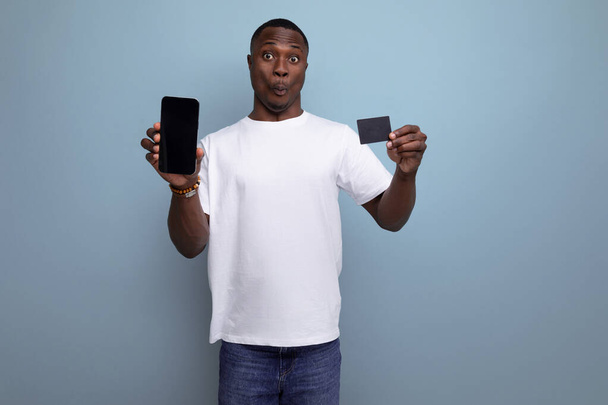 30 letý americký muž dospělý manažer v bílém tričku zobrazující displej smartphonu a nákupní kartu. - Fotografie, Obrázek