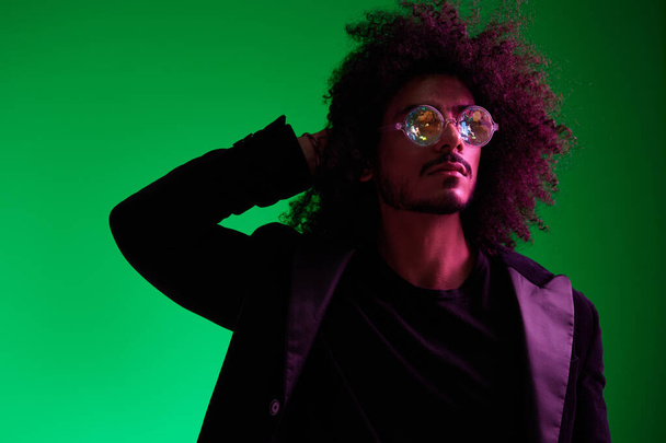 Divat portré egy férfiról göndör hajjal, zöld háttérrel, napszemüveggel, multinacionális, színes rózsaszín fénnyel, trendi, modern koncepcióval. Kiváló minőségű fénykép - Fotó, kép