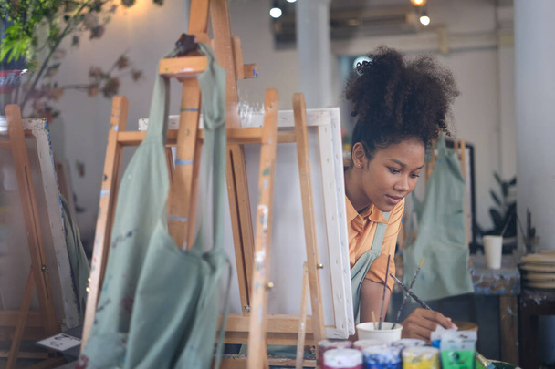 Młoda Afroamerykanka artystka mieszanie kolor obraz olejny do dzieła sztuki, siedzi przed sztalugami w studio sztuki. - Zdjęcie, obraz