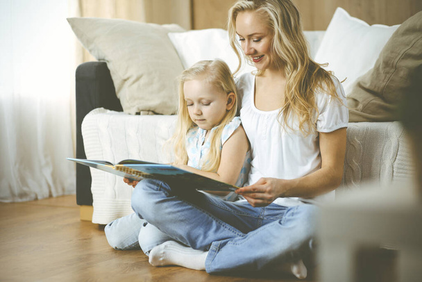 Happy family. Blonde young mother reading a book to her cute daughter while sitting at wooden floor. Motherhood concept. - Fotografie, Obrázek