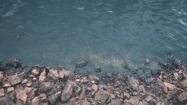 Blick von oben auf das felsige Ufer mit vielen Steinen unterschiedlicher Form, kleine Wellen auf der Wasseroberfläche. - Filmmaterial, Video