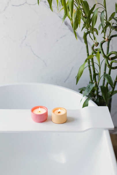 Kézműves rózsaszín és narancssárga szója gyertyák fürdőkádban közelében bambusz növény fehér fény fürdőszoba belső - Fotó, kép