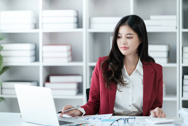 Beautiful asian businesswoman working on laptop and calculating with financial calculator from statistics data graphs, charts. Successful business results in modern office wear red shirt. High quality - Photo, Image