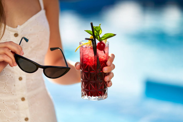 Close-up of woman hands with cocktail glass and sunglasses. Female holding a drink and sun shades near swimming pool with blue water. Girl chilling with beverage in a tropical sun. Vacation concept - Foto, Imagen