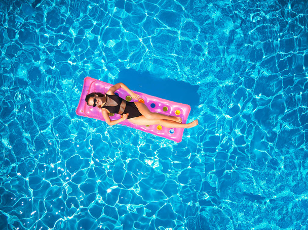 Fit pretty girl in bikini chilling on inflatable pink mattress in swimming pool. Slim hot woman in swimwear tanning. Female relaxing on a float in blue water at luxury resort. Aerial, view from above. - Foto, Imagen