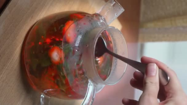 Process of brewing tea, tea ceremony, freshly brewed fruit and herbal tea. Spoonful stirs tea in teapot with tea leaves, mint, strawberries and fruit in glass teapot on table. close-up. - Footage, Video