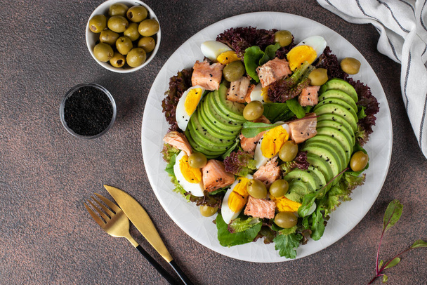 Salad with baked salmon, avocado, eggs and olives, sprinkled with sesame seeds on a white plate, Top view - Zdjęcie, obraz