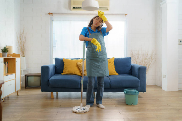 Erschöpftes asiatisches Dienstmädchen kämpft mit Müdigkeit, während sie das Haus wischt, und zeigt die Herausforderungen und harte Arbeit hinter dem Haus - Foto, Bild