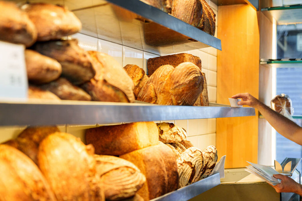 Onherkenbare werknemer in een bakkerij regelt nieuw gebakken broden en identificatiekaarten op een plank voor detailhandelsdoeleinden. - Foto, afbeelding