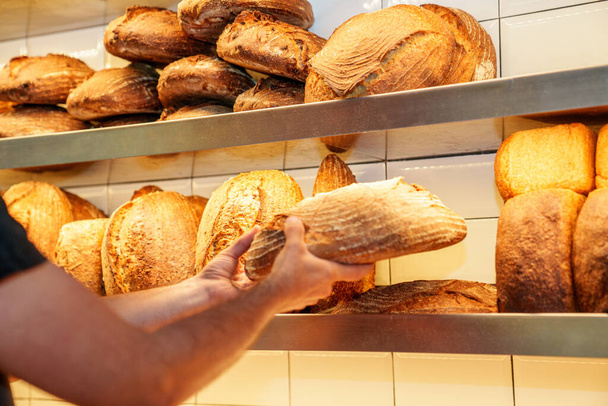 An anonymous laborer in a bakery delicately positions recently baked bread loaves onto a shelf, preparing them for sale. - Photo, image