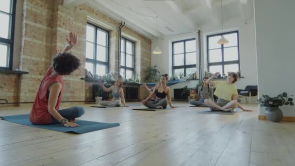 Ampio colpo di gruppo di donne in abbigliamento sportivo seduto con le gambe incrociate su stuoie e facendo curva laterale durante la pratica dello yoga con istruttrice donna - Filmati, video