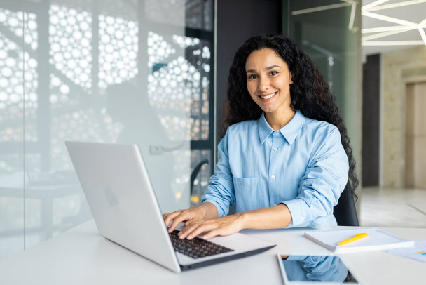 Young beautiful Indian female programmer working inside the office on a laptop, woman smiling and looking at the camera, portrait of a satisfied and confident businesswoman at the workplace. - Zdjęcie, obraz