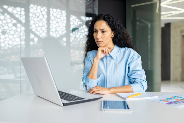 Serious thinking woman working inside office at workplace with laptop, confident hispanic woman looking at technical task, brainstorming solution, business woman with curly hair looking at laptop - Fotoğraf, Görsel