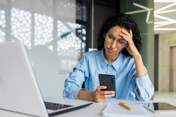 Shocked and upset business woman working inside office at workplace, depressed hispanic woman reading bad news using app on smartphone, holding phone. - Foto, imagen