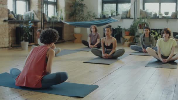 Grupo de chicas deportistas practicando estiramiento de mariposa en esteras en estudio durante clase de yoga con profesora - Metraje, vídeo