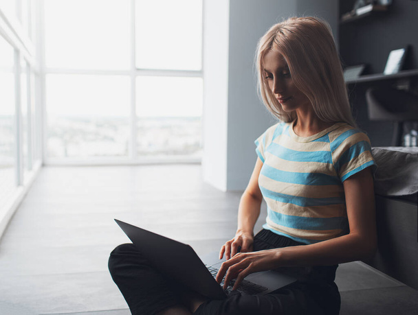 Портрет блондинки, сидящей на полу при использовании ноутбука на фоне панорамных окон квартиры. - Фото, изображение