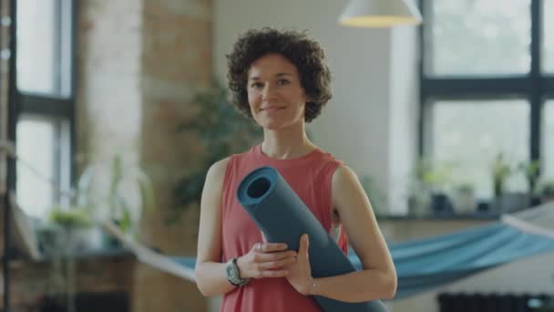 Waist up tracking shot of cheerful woman in sportswear holding exercise mat and posing for camera with smile in yoga studio - Footage, Video