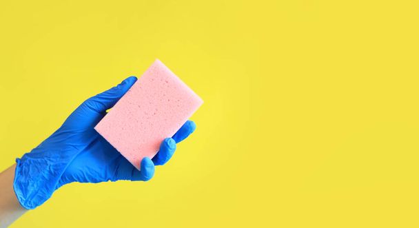 青い手袋の女性の手は皿を洗うためのスポンジを保持します。掃除だ。明るい黄色の背景にピンクのスポンジで手。広告のクリーニング - 写真・画像