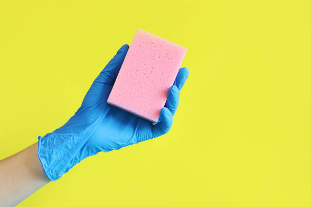 A mão de uma mulher em uma luva azul segura uma esponja para lavar pratos. Limpeza da casa. Uma mão com uma esponja rosa sobre um fundo amarelo brilhante. Publicidade de limpeza - Foto, Imagem