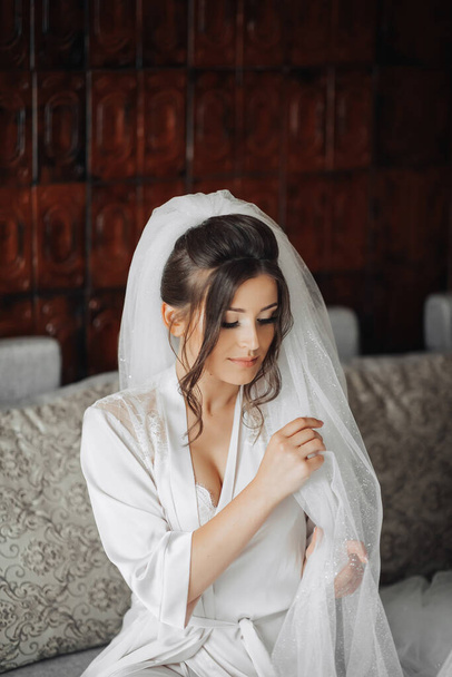 Portrait of the bride. A brunette bride is sitting on a gray sofa in a dressing gown, posing, holding her voluminous white veil. Gorgeous make-up and hair. Wedding photo. Beautiful bride - Fotó, kép